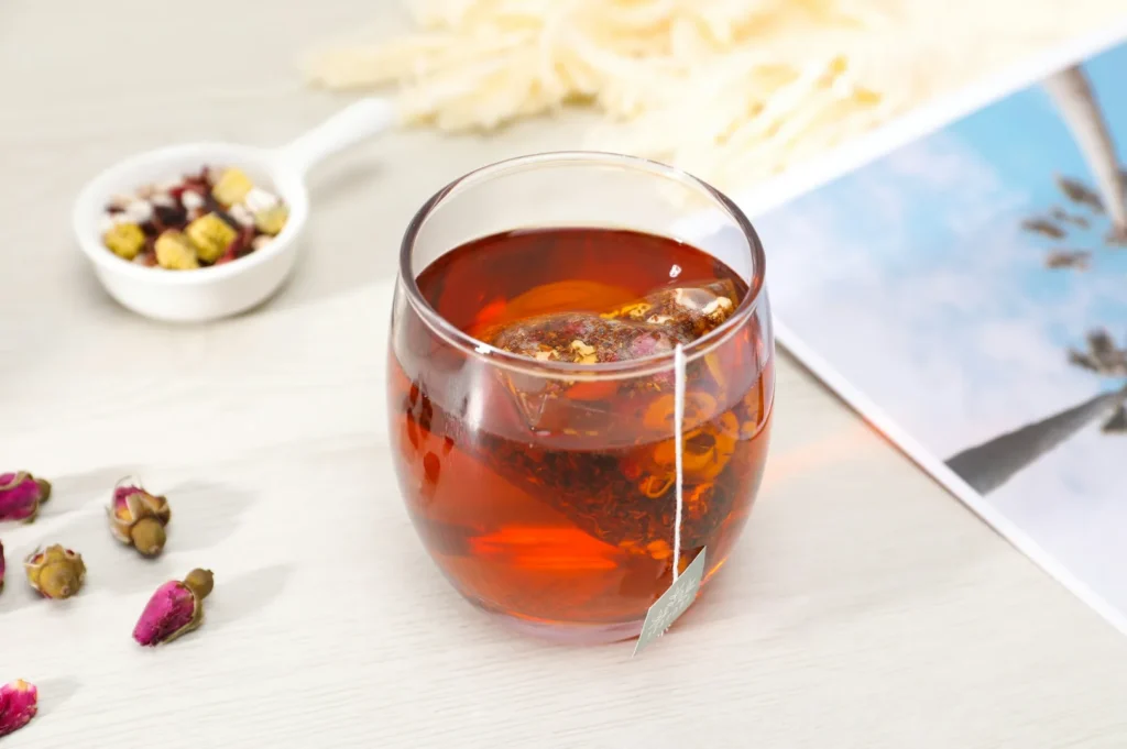 A glass of red herbal tea with a tea bag on a light wooden table, surrounded by dried rosebuds and a small bowl of mixed dried flowers, creating a calming and cozy atmosphere.