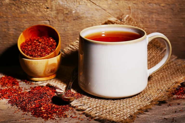 A white ceramic mug filled with reddish-brown tea sits on a rustic wooden surface, surrounded by loose tea leaves, a small wooden bowl, and a spoon on a burlap cloth.