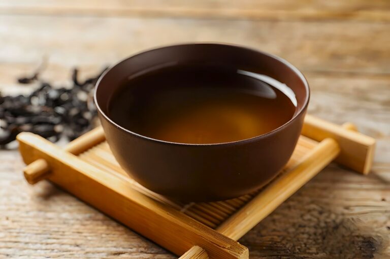 A minimalist tea setting featuring a matte brown cup on a wooden rack, the simmering amber liquid reflects light, with distant, unfocused tea leaves alluding to Wuyi oolong craftsmanship.