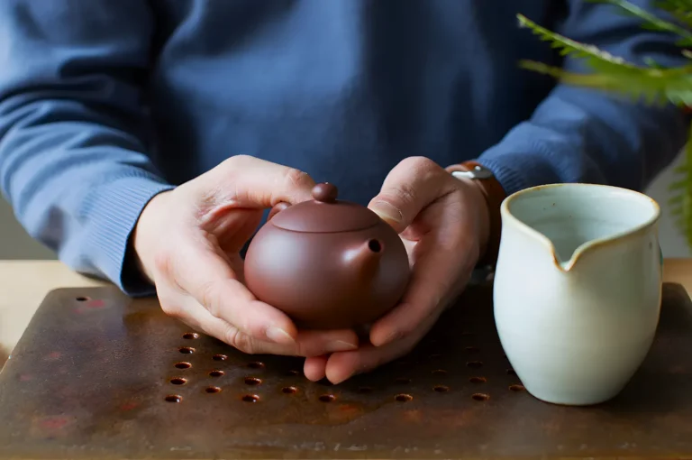 A person in a blue sweater holds a small clay teapot over a wooden tray, next to a white ceramic pitcher. The scene conveys a calm, meditative tone.