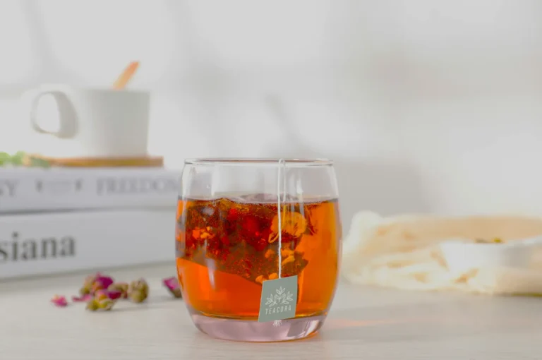 A glass filled with rich amber tea from a Teabags sachet sits on a table. Nearby, closed books and dried flowers create a calm, cozy atmosphere.