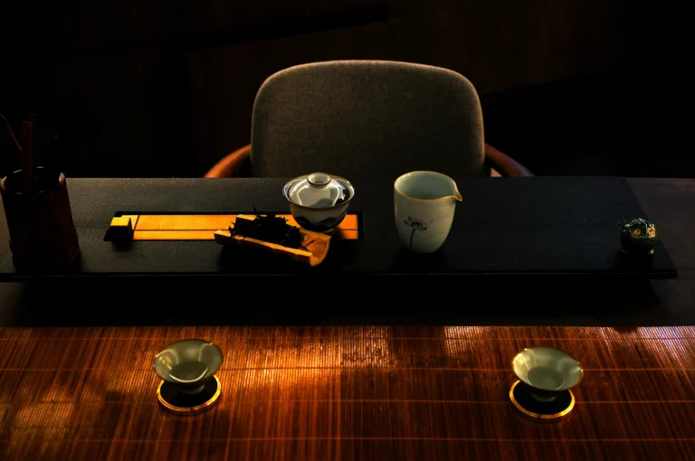 Dimly lit tea ceremony setup with a teacup, teapot, and small bowls on a wooden table. A calm, serene atmosphere is created by the minimalist arrangement.