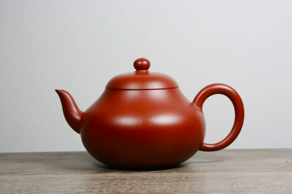 A smooth, reddish-brown teapot with a round body, curved spout, and loop handle sits on a wooden surface against a plain white background.