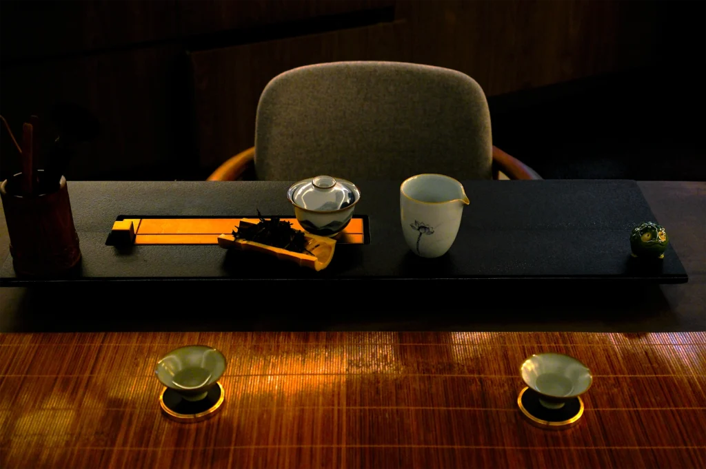 A dimly lit tea ceremony setup with a black tray holding a teapot, cup, and tea leaves. Two small bowls rest on a wooden table, creating a serene atmosphere.