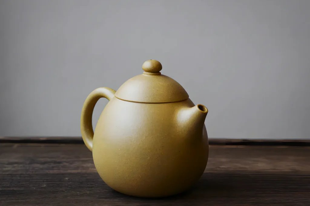 A simple brown teapot with a round body and curved handle sits on a wooden surface against a plain gray background, conveying a minimalist and calm tone.