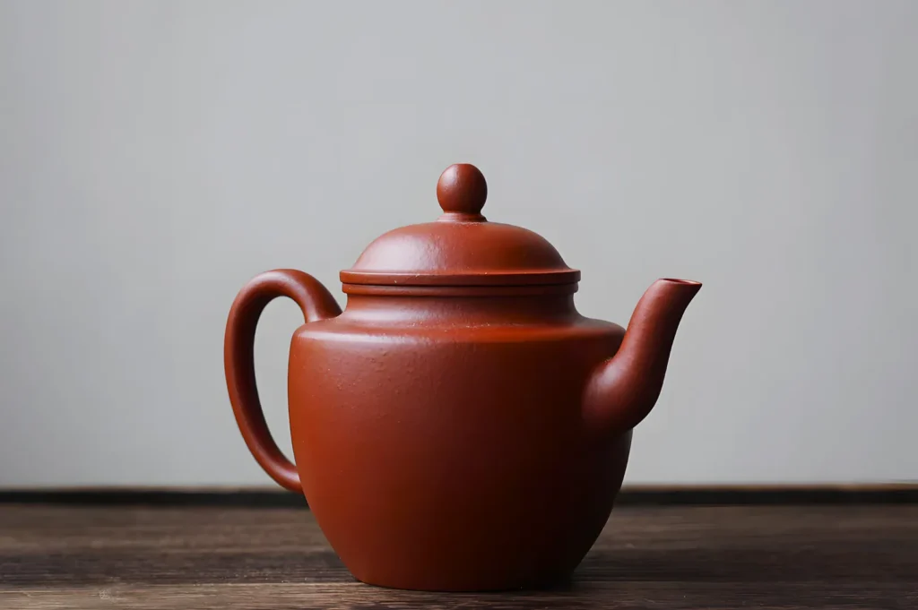 A smooth, brown ceramic teapot with a round lid sits on a wooden surface. The background is a neutral gray, creating a minimalist, serene tone.