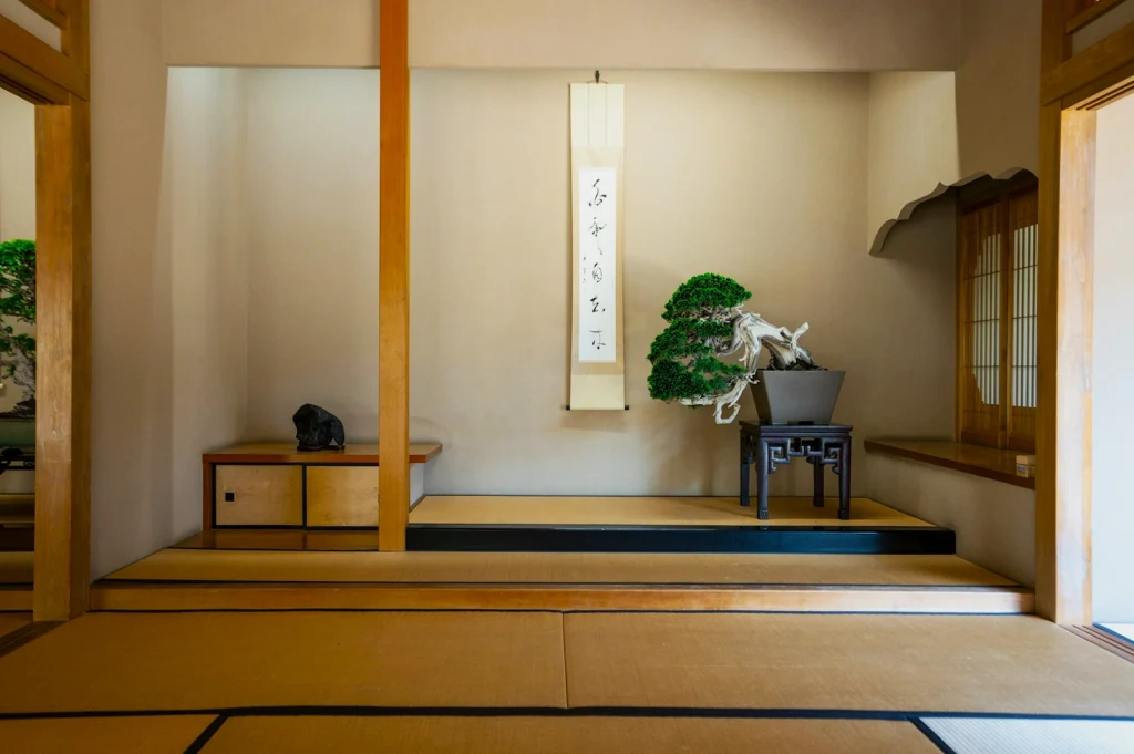 Tranquil Japanese room with tatami mats, a bonsai tree on a wooden table, and a scroll with calligraphy on the wall. Minimalist and serene ambiance.