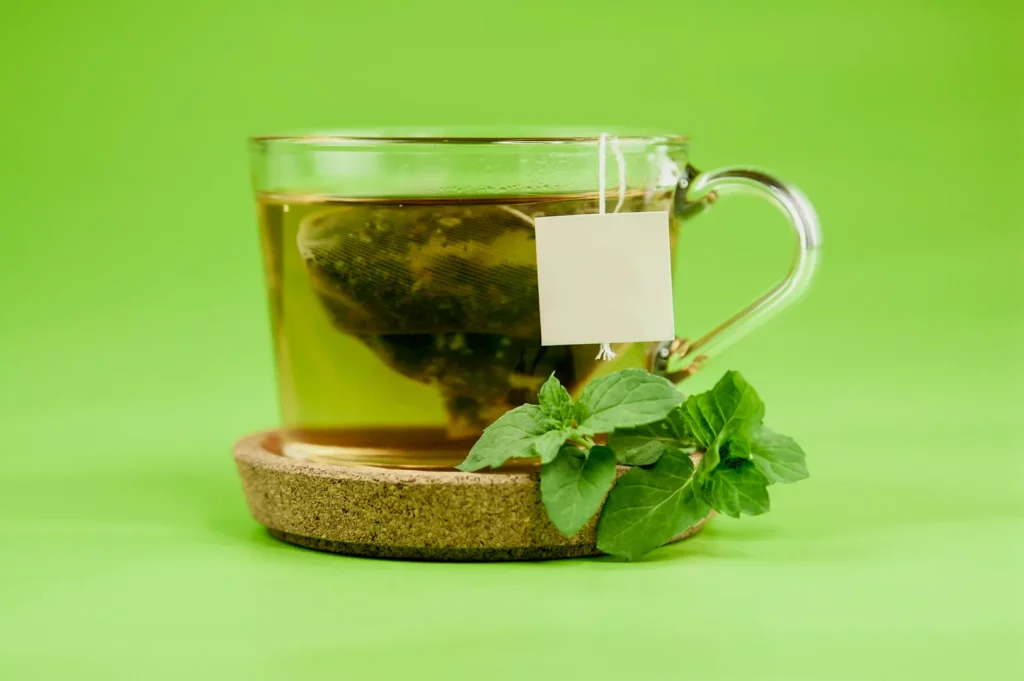 Glass cup of tea with a teabag sits on a cork coaster against a green background. Fresh mint leaves are placed beside the cup, conveying a refreshing feel.