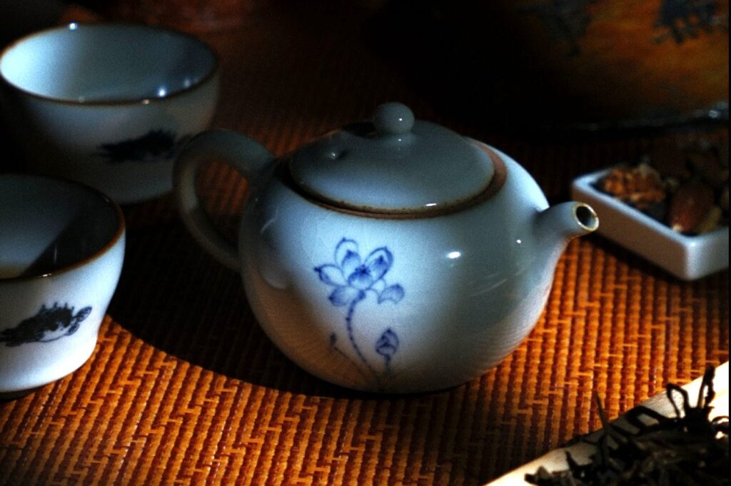 A delicate white teapot with blue floral designs sits on a woven mat, surrounded by two matching cups and a dish of nuts, creating a warm, peaceful atmosphere.