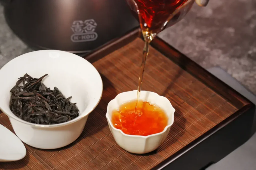 Pouring vibrant amber tea into a small white cup on a wooden tray, next to a bowl of loose leaf tea. Warm, inviting atmosphere.