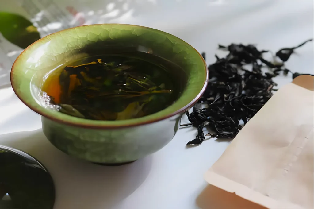 A green cup filled with brewed tea sits on a white surface, surrounded by scattered dried tea leaves and an open tea packet, under soft lighting.