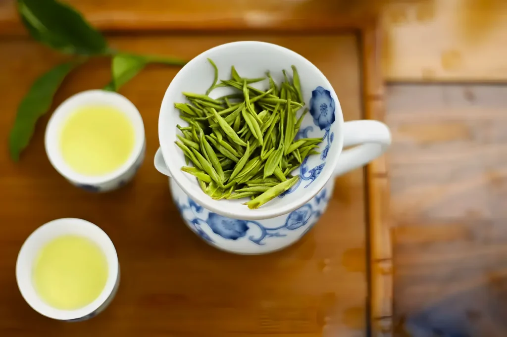 A ceramic cup with blue floral patterns holds fresh green tea leaves. Two cups of yellow-green tea sit beside it on a wooden tray, creating a calm, inviting scene.