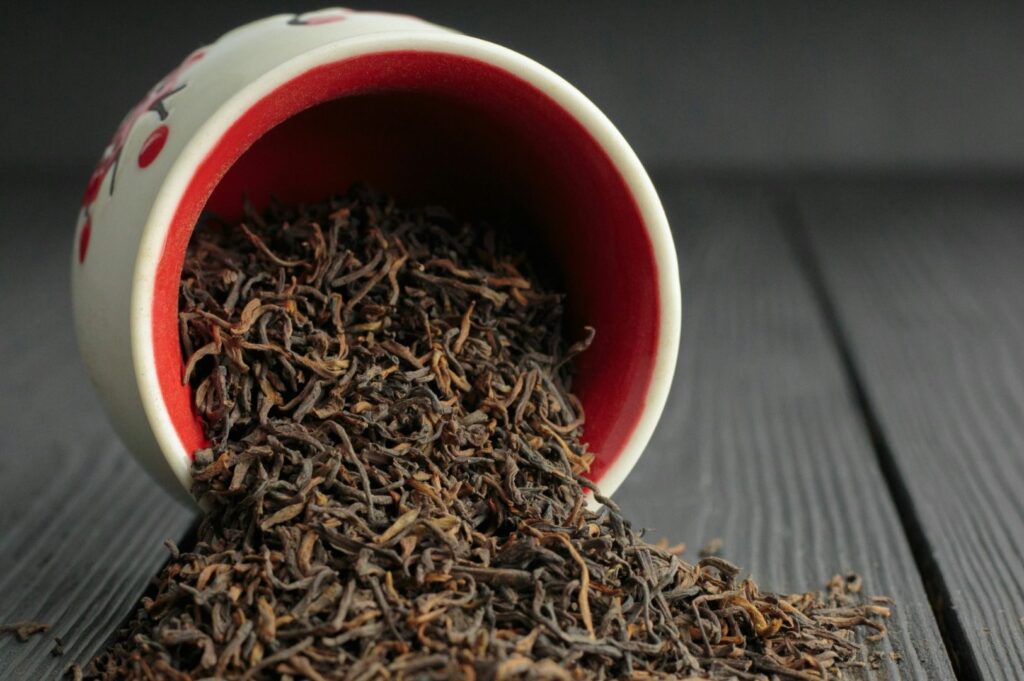 A ceramic cup with a red interior lies tipped on its side, spilling dark, twisted loose tea leaves onto a weathered wooden surface, showcasing rich browns and varied textures.