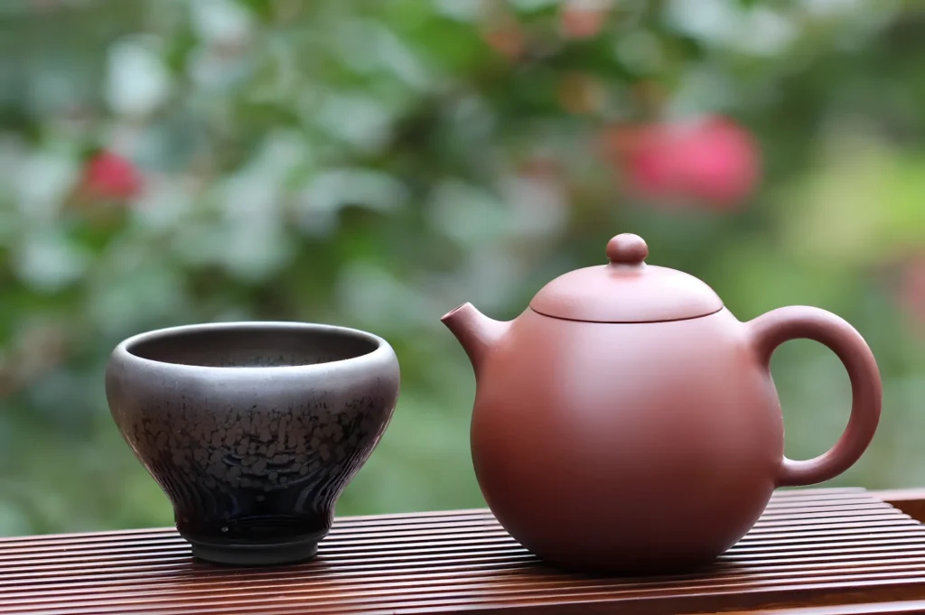 A brown teapot and a textured ceramic cup sit on a wooden surface. The background features a garden with blurred green foliage and pink flowers.