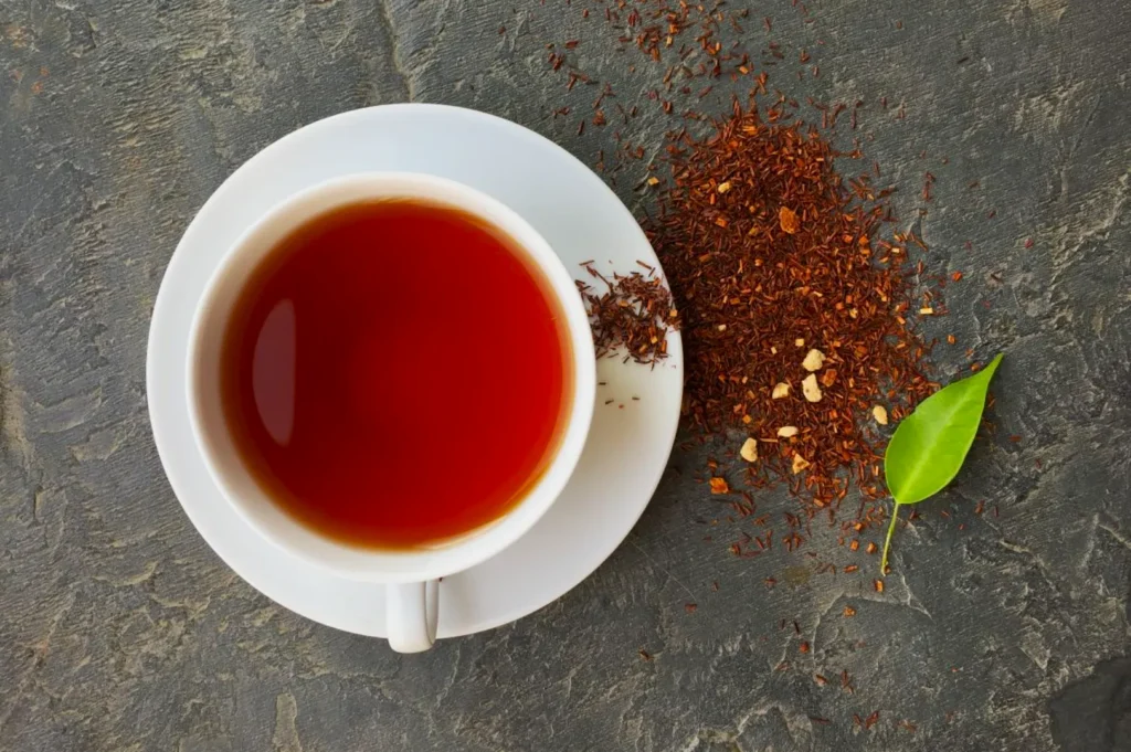 A white cup filled with red rooibos tea sits on a saucer. Loose rooibos leaves and a green leaf are scattered on the textured gray surface.