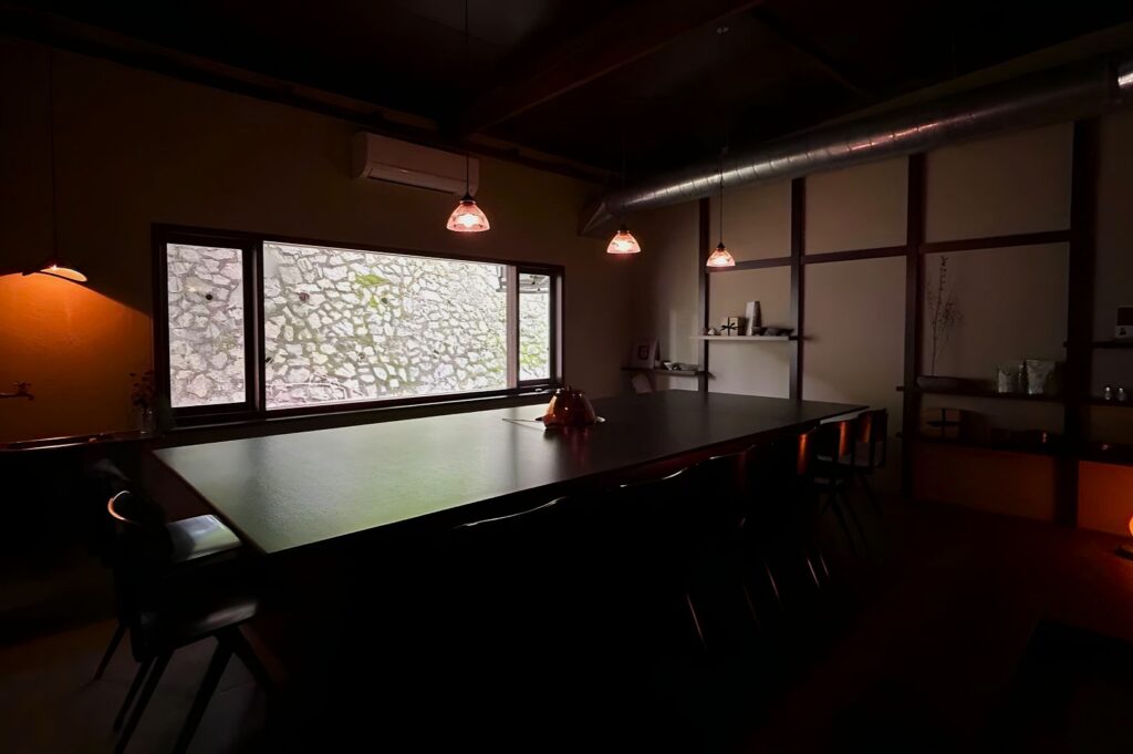 A professional still-life shot featuring a glossy brown tea bowl on a wooden rack, its steaming surface catching light while distant, out-of-focus Wuyi oolong leaves hint at the tea's origin.