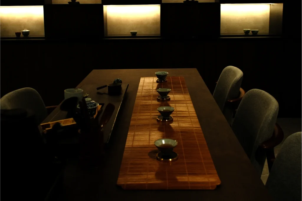 Dimly lit tea room with a wooden table featuring a bamboo mat and six small teacups. Warm lights illuminate shelves in the background, creating a serene atmosphere.