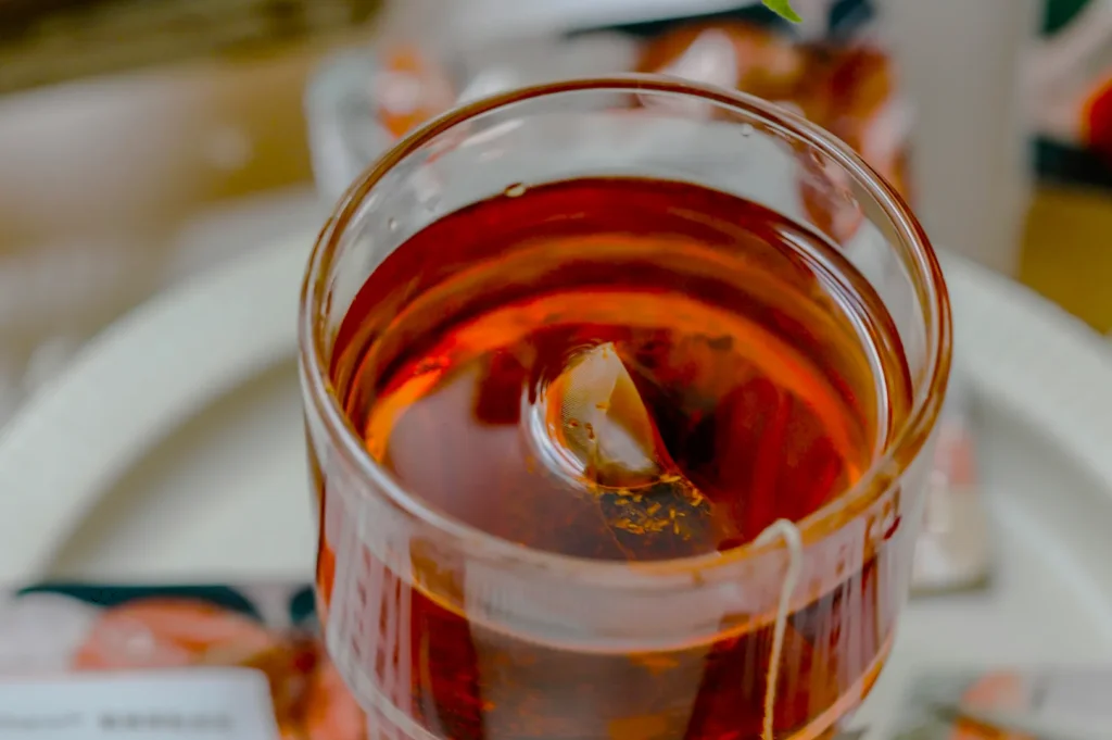 Close-up of a glass of iced tea with a tea bag and ice cube, sitting on a white plate. The tea is a rich amber color, evoking freshness.
