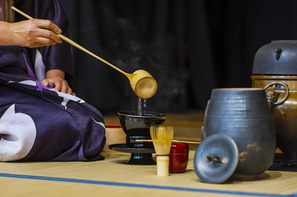 A person in a kimono carefully pours hot water using a bamboo ladle into a black teacup, surrounded by traditional tea ceremony utensils on a tatami mat.