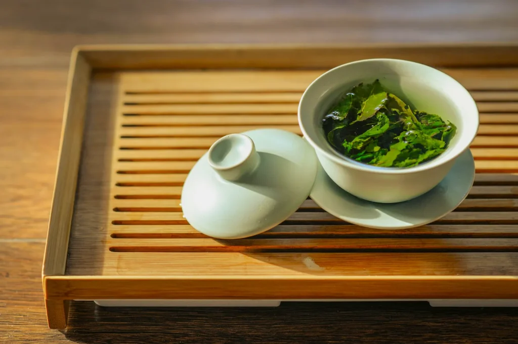 A white ceramic gaiwan with green tea leaves is placed on a wooden slatted tray. The scene is sunlit, evoking a calm and serene atmosphere.