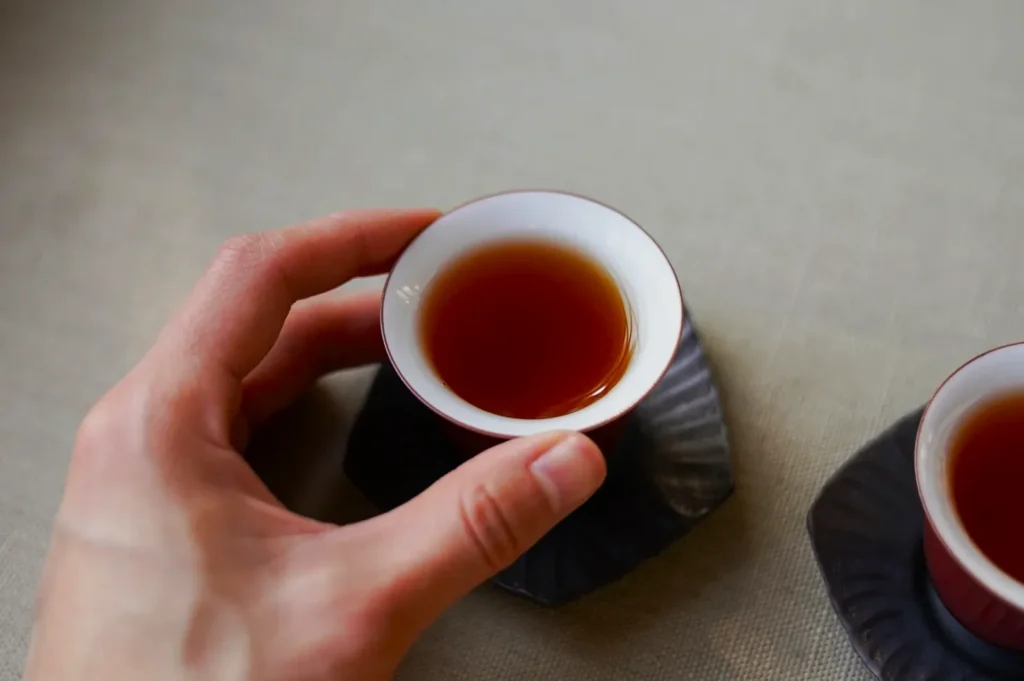 A hand holding a small cup filled with brown tea, resting on a textured saucer. Another similar cup is partially visible on a neutral tablecloth background.