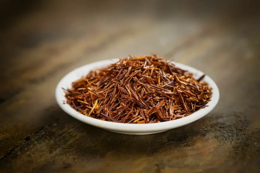 A white bowl filled with dried, red rooibos tea leaves sits on a rustic wooden surface. The scene conveys a natural, earthy tone.