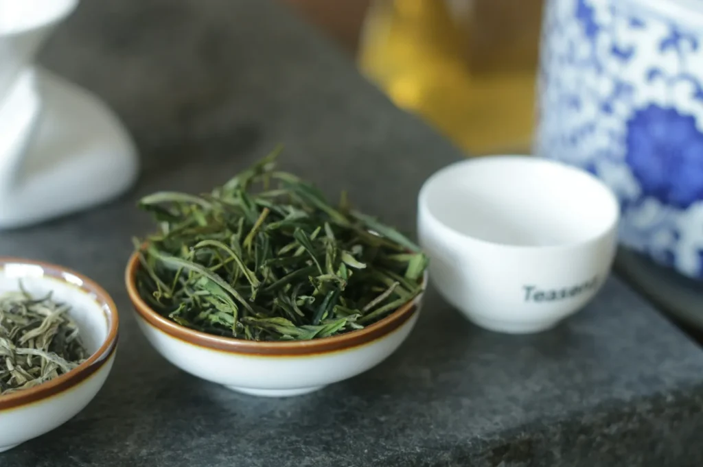 A small white bowl filled with green tea leaves sits on a dark surface. Beside it, an empty white teacup and a partially visible porcelain jar with blue patterns convey a calm, traditional tea setting.