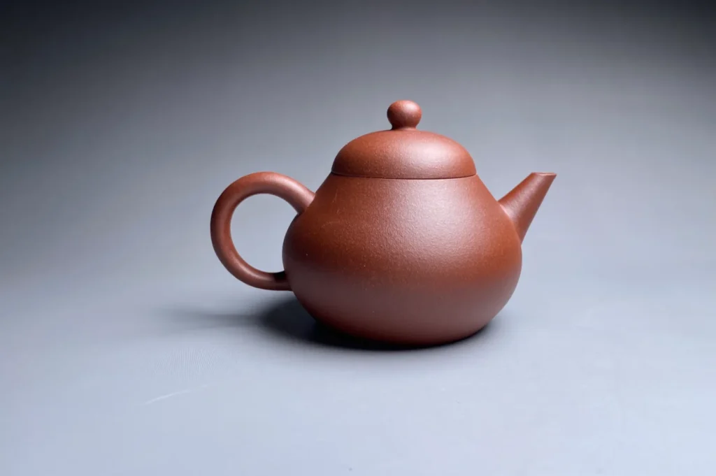 A brown clay teapot with a smooth, rounded shape sits on a gray surface. The lighting highlights its simple elegance, evoking a calm, serene atmosphere.