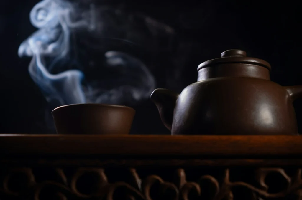 Dark, atmospheric scene of a steaming teapot and cup on an ornate wooden surface. Swirling steam adds a sense of warmth and tranquility.