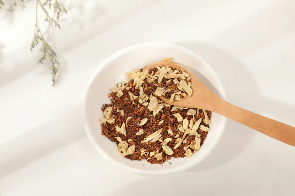 A white bowl filled with loose tea leaves and flower petals is accompanied by a wooden spoon. The setting is bright, with soft lighting and a floral accent.