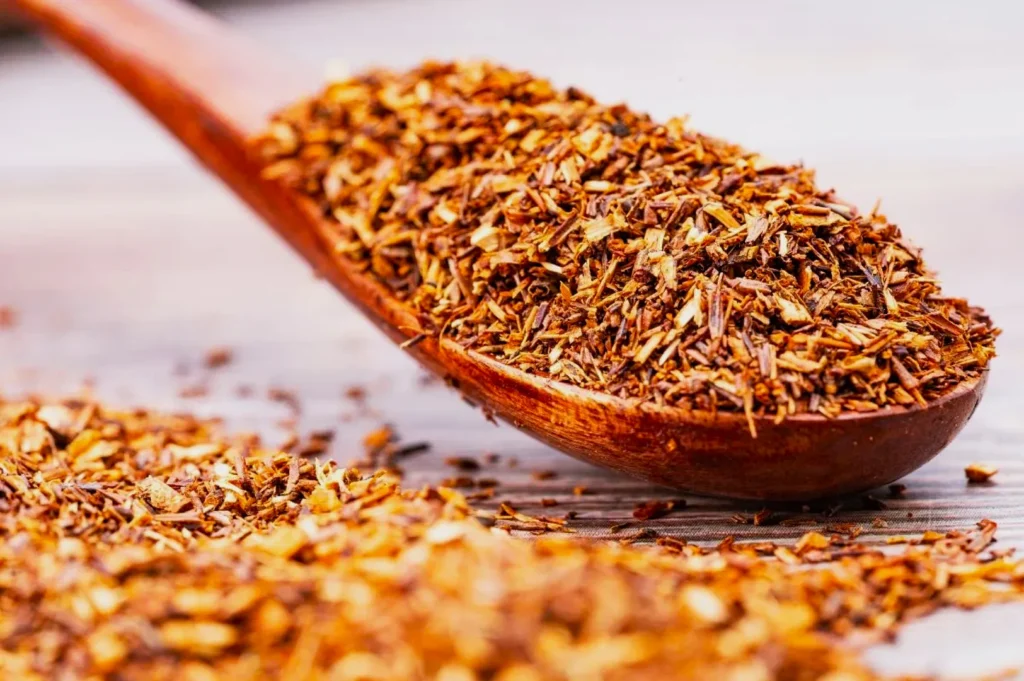 Close-up of a wooden spoon filled with loose rooibos tea leaves on a wooden surface. The brown, dried leaves create a warm, rustic feel.