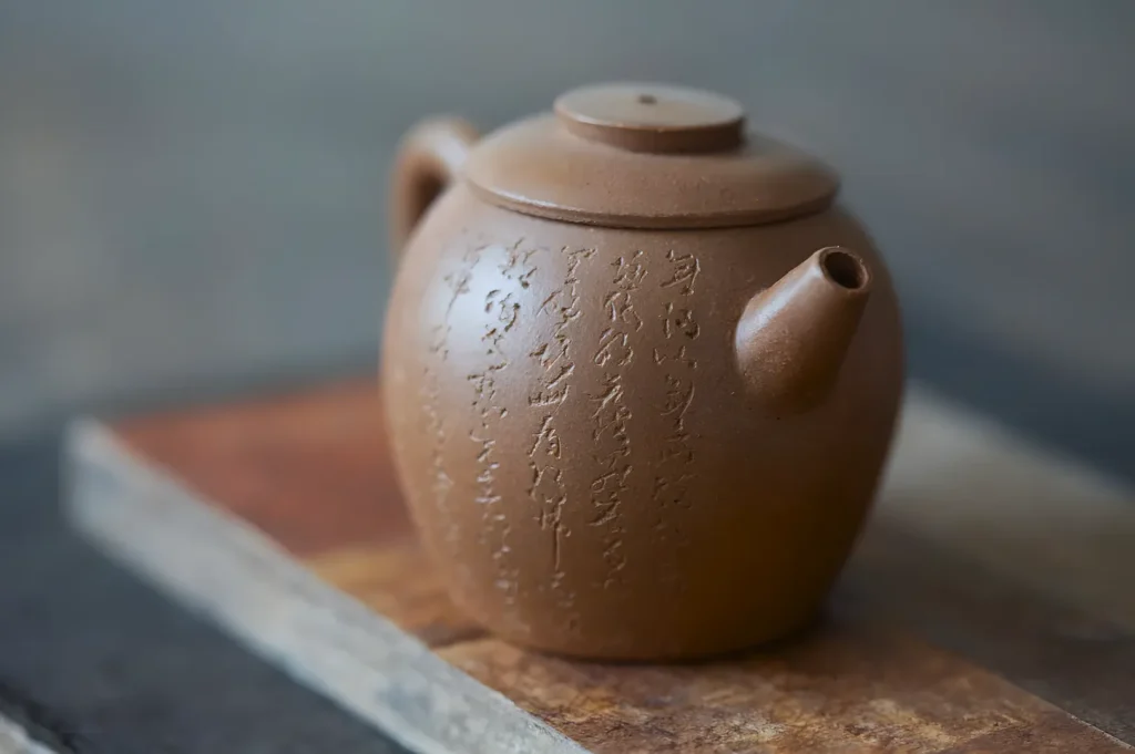 A brown clay teapot with intricate engraved characters sits on a wooden board. The teapot's round shape and earthy tone convey a rustic elegance.