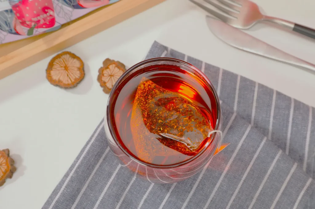 A glass of red tea with a teabag on a striped cloth. Dried citrus slices are nearby, along with a fork and knife. Warm and inviting setting.
