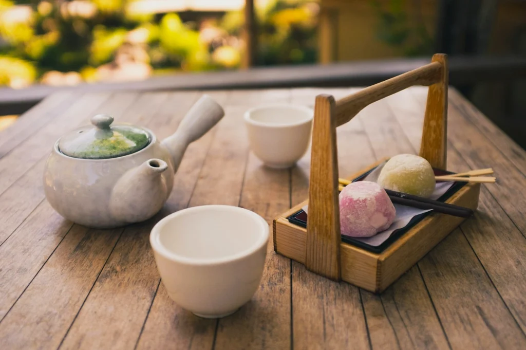 A serene tea setting on a wooden table includes a white teapot, two cups, and a wooden tray with pink and white mochi, creating a peaceful, inviting atmosphere.