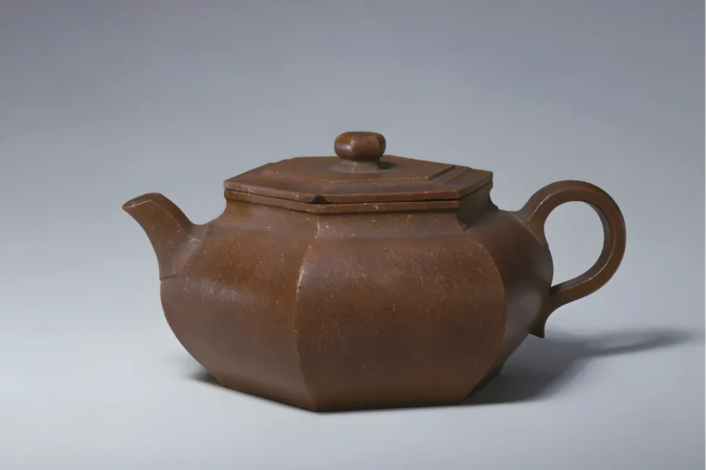 A brown, hexagonal teapot with a curved spout and a loop handle, set against a plain gray background. The design is simple, conveying rustic elegance.
