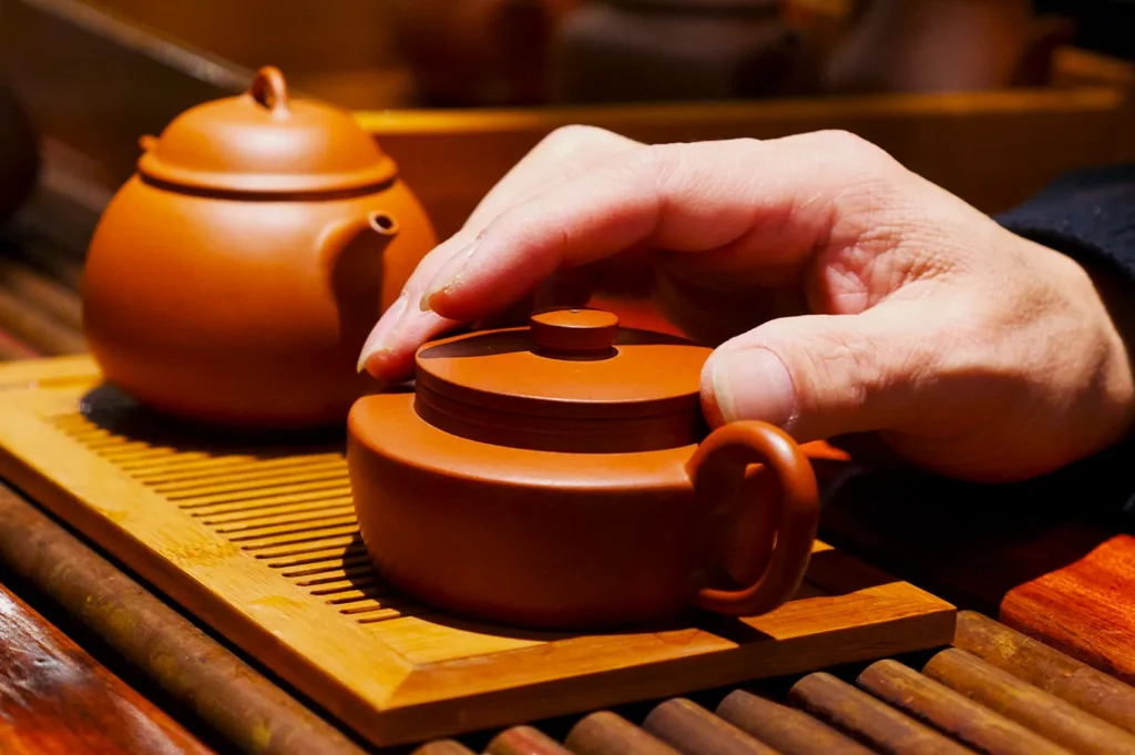 A hand gently places a lid on a small, brown clay teapot on a bamboo mat. Another teapot is nearby, creating a serene, traditional tea setting.