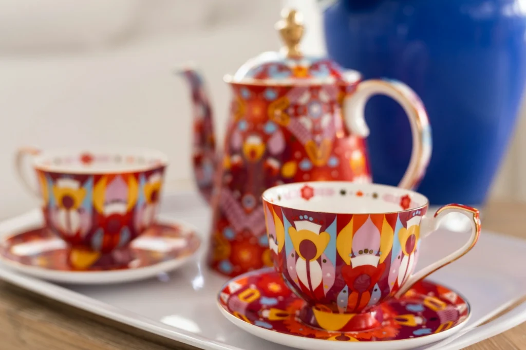 Vibrant tea set with floral patterns on red cups and teapot, placed on a tray. Warm, inviting elegance against a blurred blue vase backdrop.
