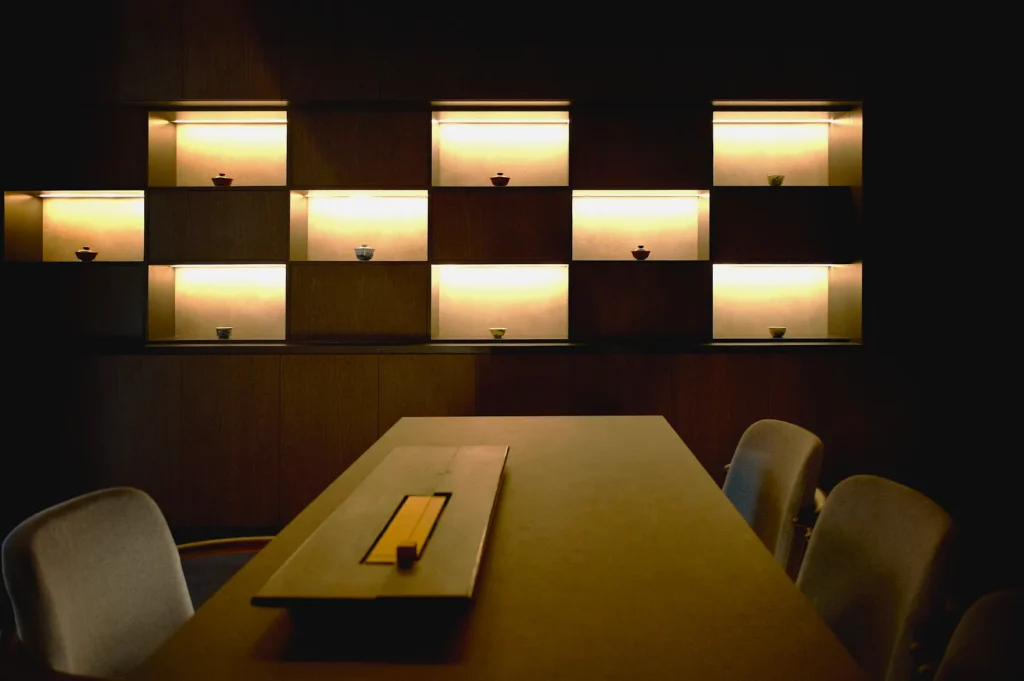 A dimly lit room with a wooden table and chairs in the foreground. Backlit shelves display small pottery, creating a serene and minimalist atmosphere.