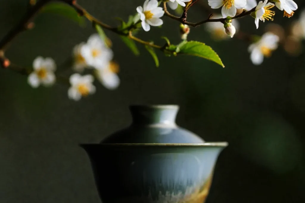 A serene scene with a green ceramic teapot under blossoming white flowers on a branch, set against a blurred dark green background, creating a tranquil and elegant mood.