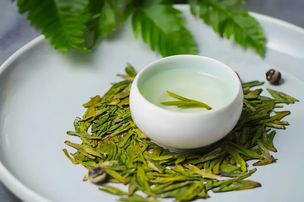 A white bowl of light green tea rests on a bed of dried green tea leaves on a circular white plate. Green leaves are blurred in the background.