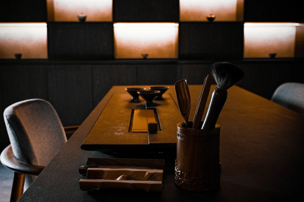 Dimly lit room with a wooden table featuring a bamboo holder of brushes and spoons. Shelves in the background have small bowls, with warm, soft lighting.
