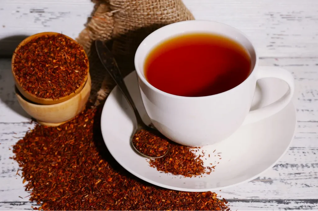 A white cup filled with reddish-brown rooibos tea sits on a saucer, surrounded by loose rooibos leaves and a wooden container, evoking warmth and coziness.