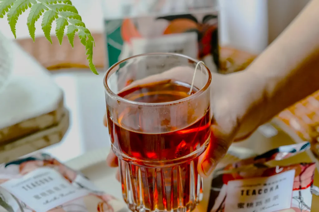 A hand grasps a glass of amber tea, evoking warmth. Green fern leaves hover above, with tea packaging and woven textures blurred in the background.