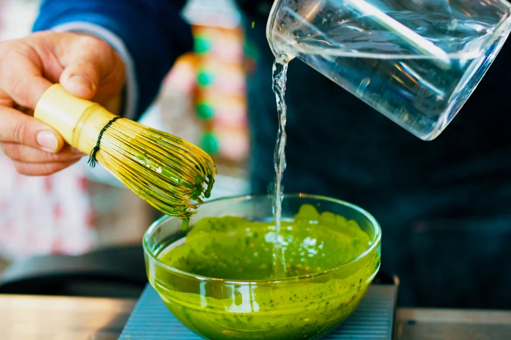 Hands prepare matcha in a bowl using a bamboo whisk, as water is poured from a pitcher. The scene emphasizes a calm, focused ritual.