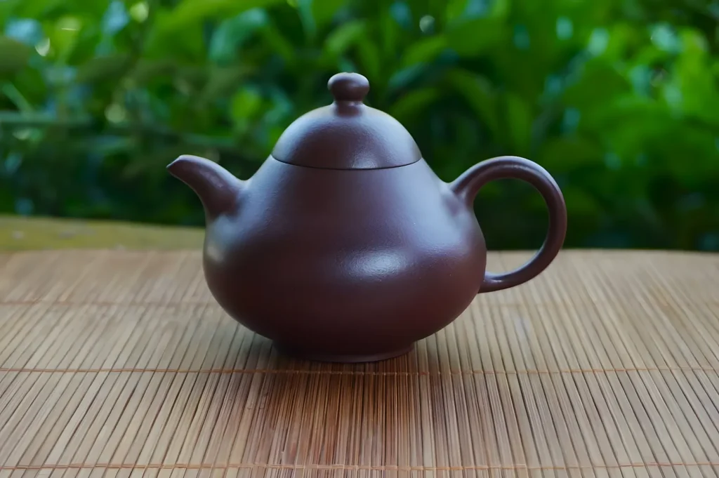 A round, brown teapot with a short spout and curved handle sits on a bamboo mat. The background showcases lush green foliage, adding a serene feel.
