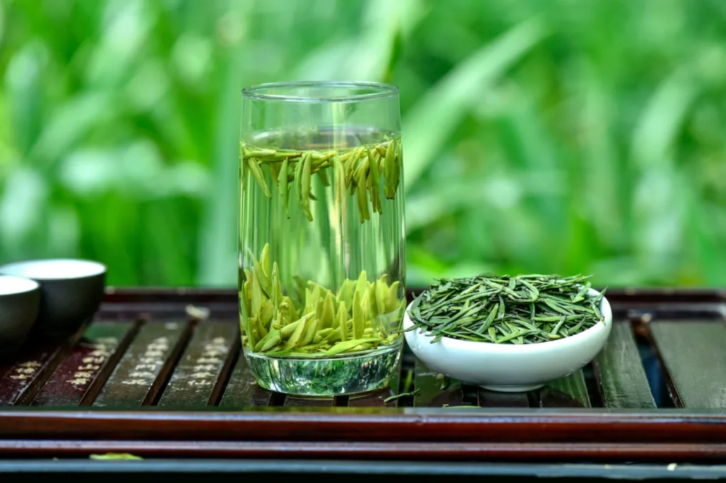 A glass filled with brewing green tea leaves on a wooden tray, next to a small white bowl of loose leaves. The background is lush greenery.