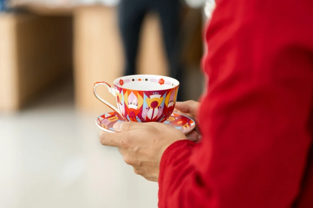 Person in a red shirt holding a vibrant, colorful teacup and saucer with abstract patterns. The setting is soft-focused, creating a warm, cozy atmosphere.