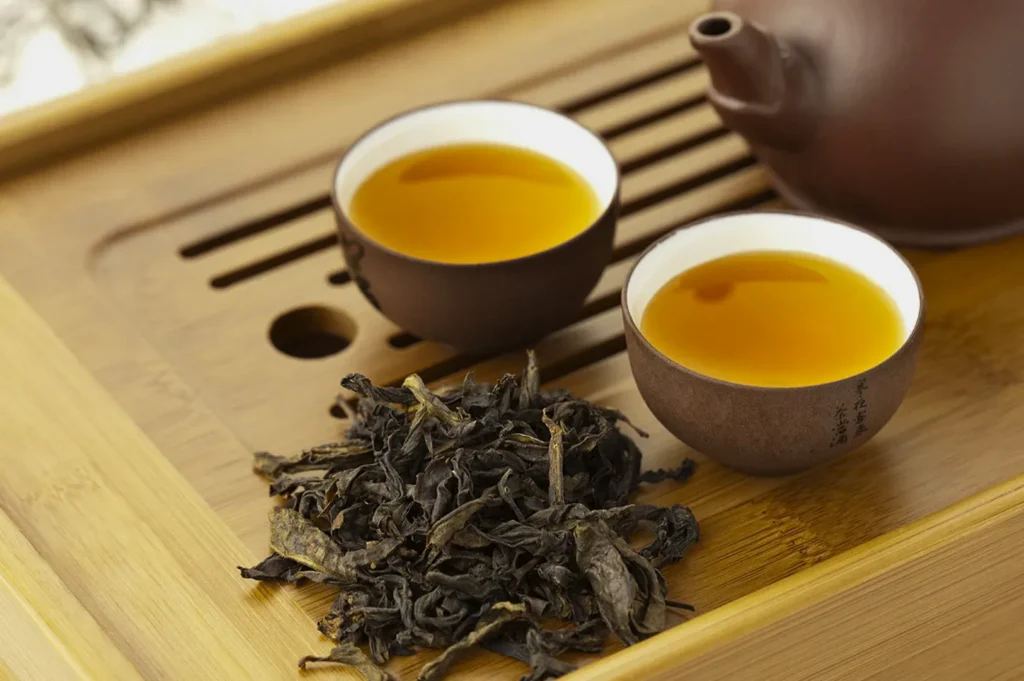 Two small cups of golden tea and loose dark tea leaves on a bamboo tray, next to a rustic teapot. The scene evokes a warm, calming atmosphere.