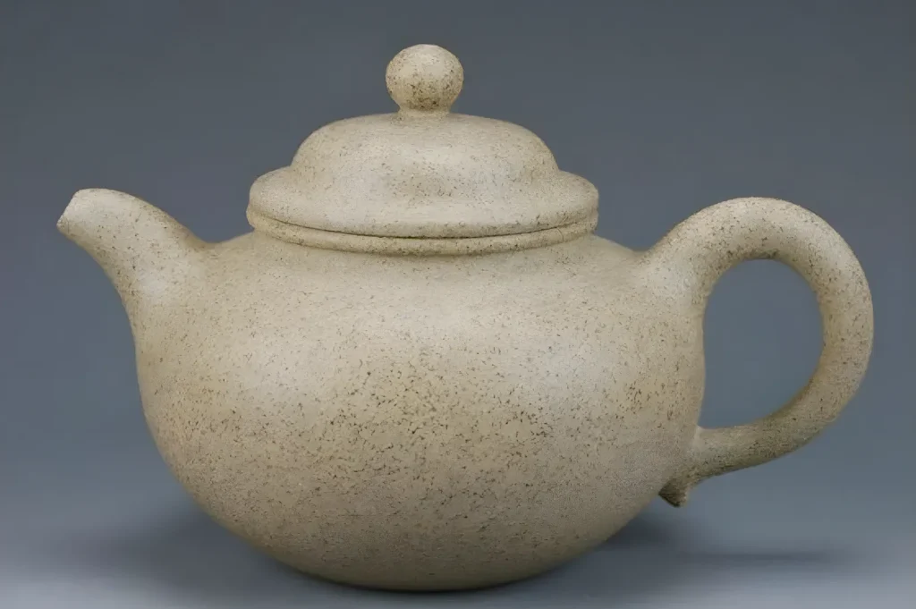 A round, speckled beige teapot with a short spout, domed lid, and curved handle sits against a gray background, conveying simplicity and elegance.