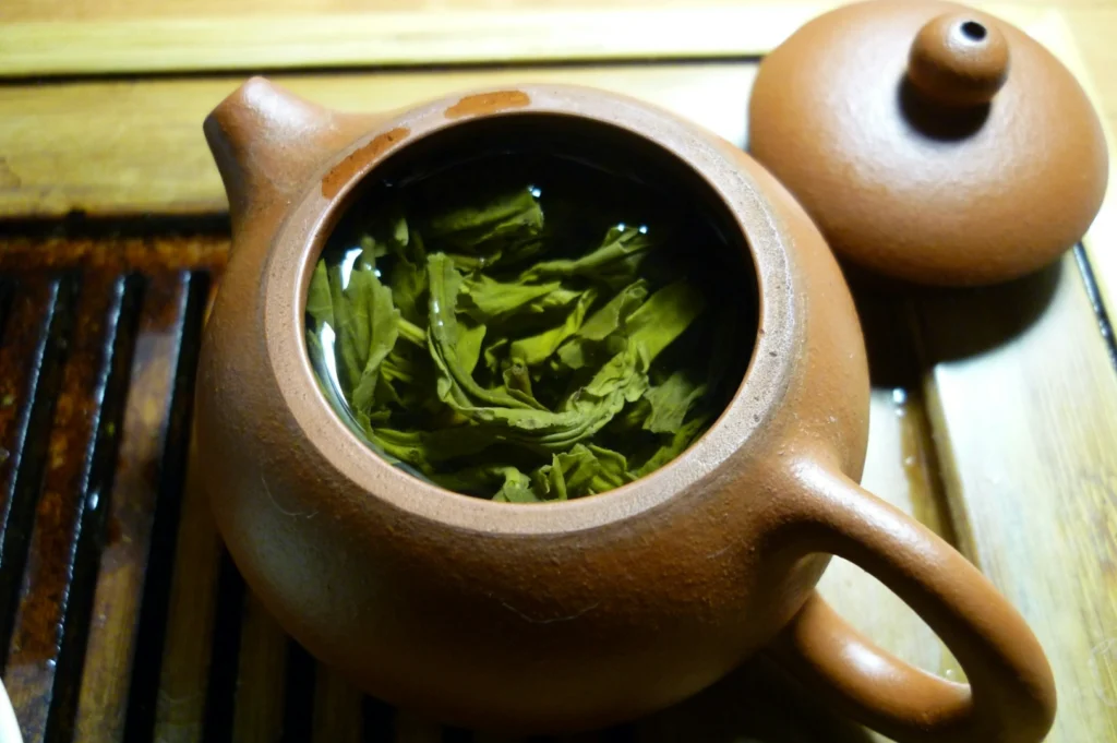 Clay teapot with green tea leaves steeping inside, lid resting beside it on a wooden surface. Warm tones suggest a cozy, tranquil scene.